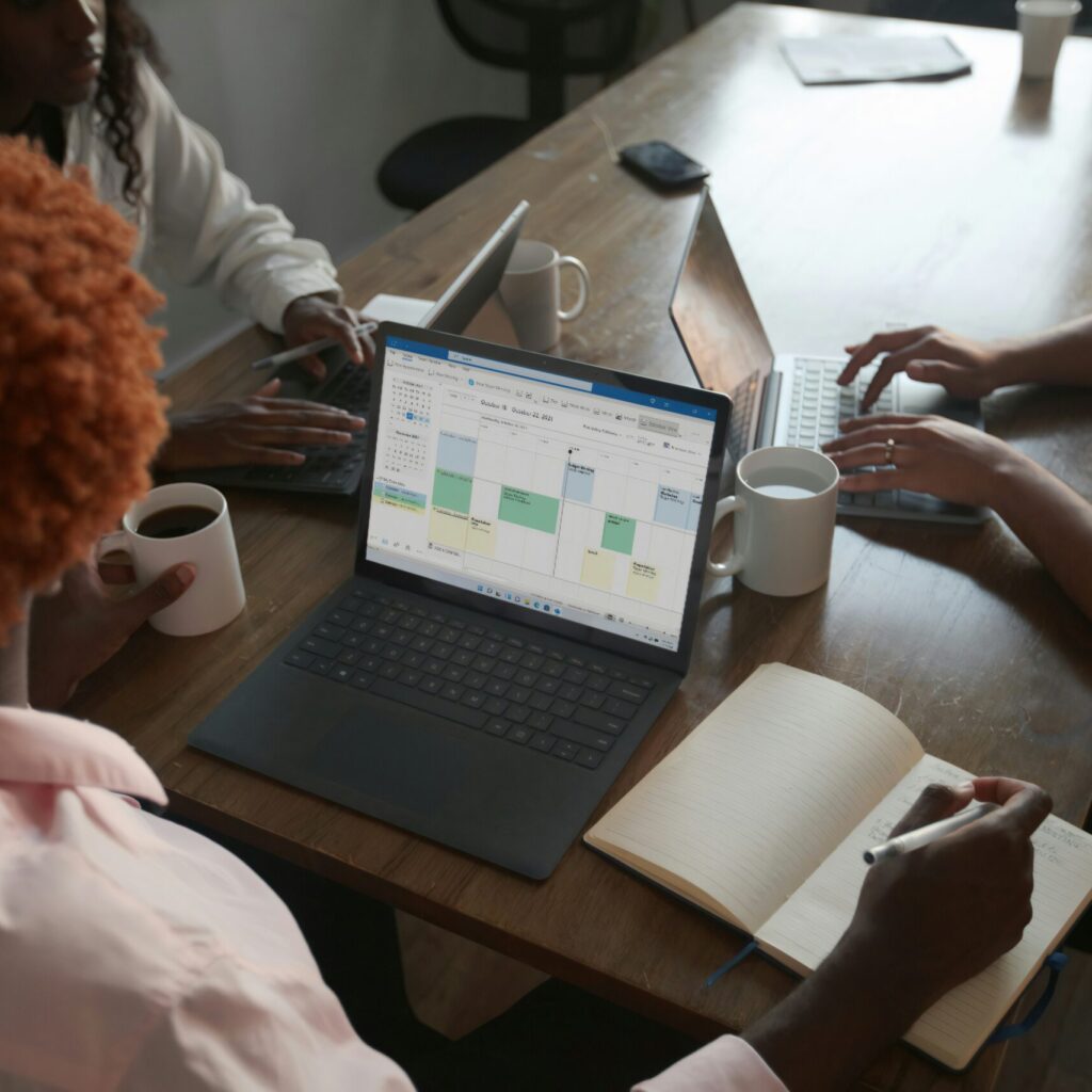 Three people in a meeting at a table discussing schedule on their Microsoft laptop